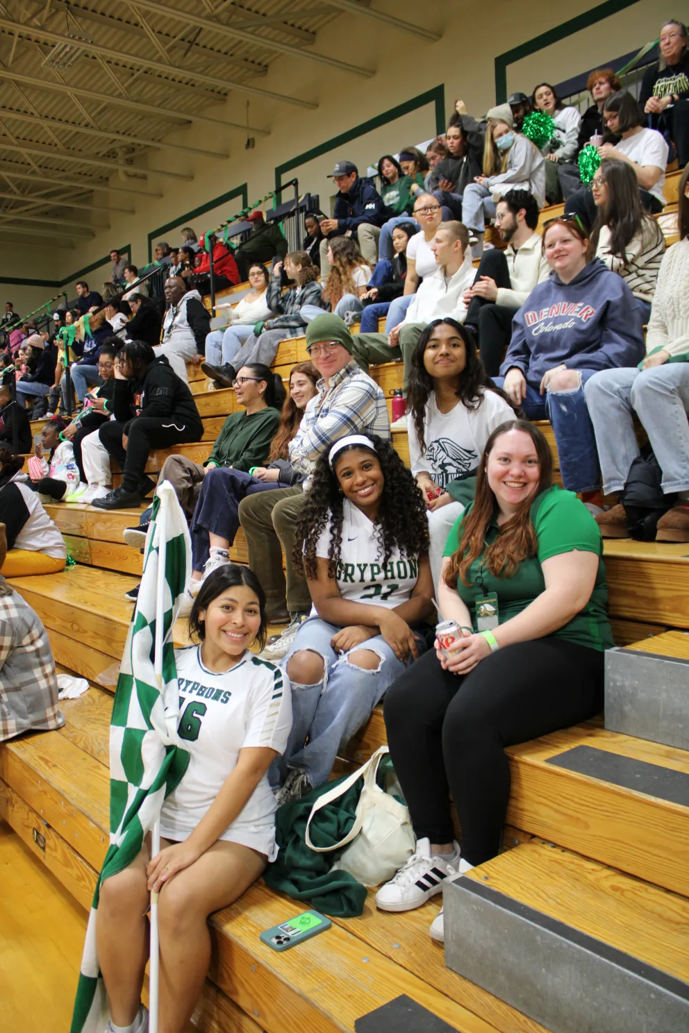 People sitting in bleachers, some smiling and wearing team shirts, with a green and white flag visible in the foreground.