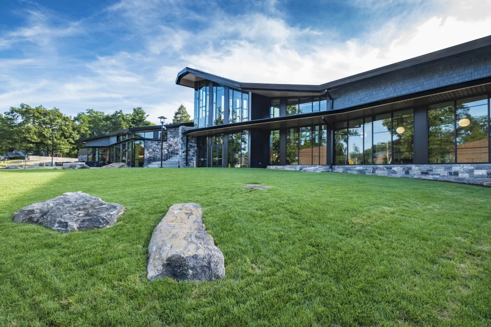 Modern building with glass facade surrounded by lush green lawn and large rocks, set against a backdrop of trees and a partly cloudy sky.