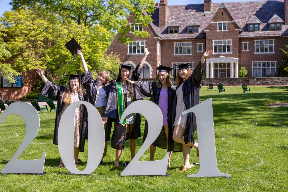 Graduates in caps and gowns celebrate by large "2021" sign on a grassy area, with a brick building and trees in the background.