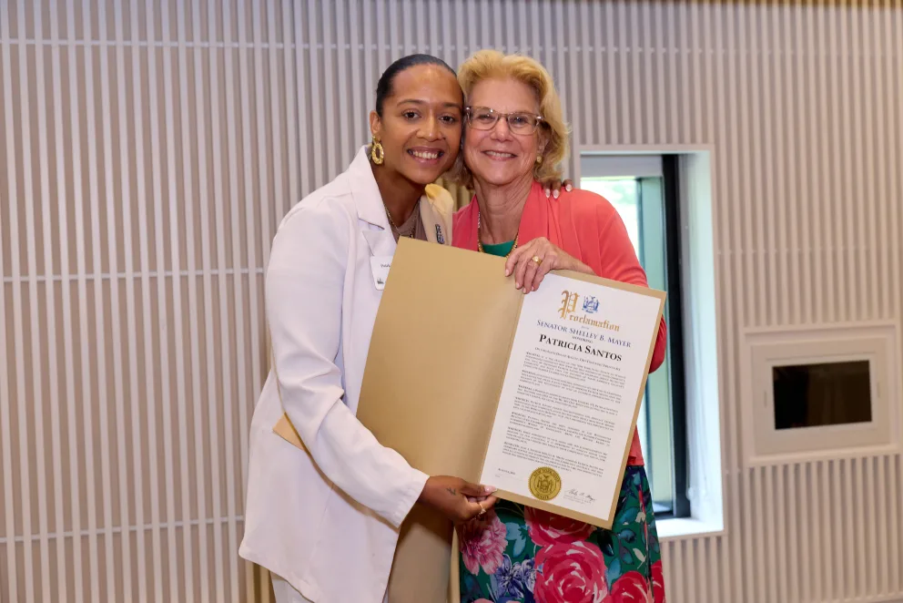 Two people smiling, holding a framed certificate together in a room with striped walls and a small window.
