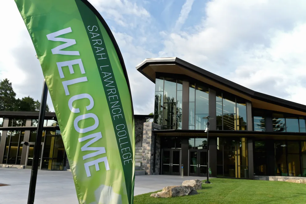 Green welcome flag for Sarah Lawrence College stands in front of modern building with large windows and stone accents under a cloudy sky.