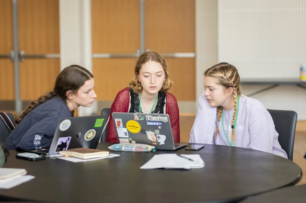Three individuals sitting at a table, focused on a laptop with stickers, surrounded by papers and notebooks in a spacious room.