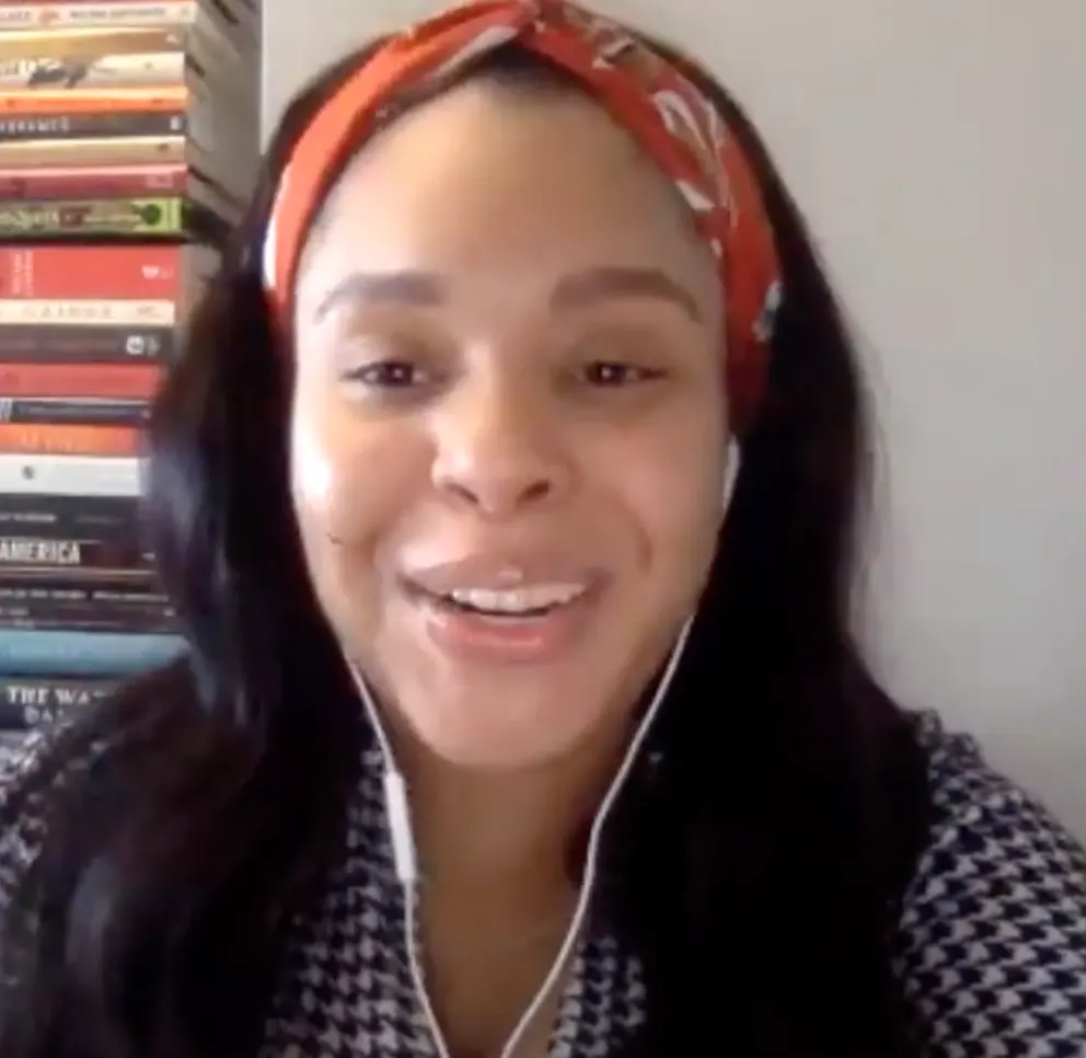 Person smiling with long hair, wearing a headband and earphones, positioned in front of a stack of books.