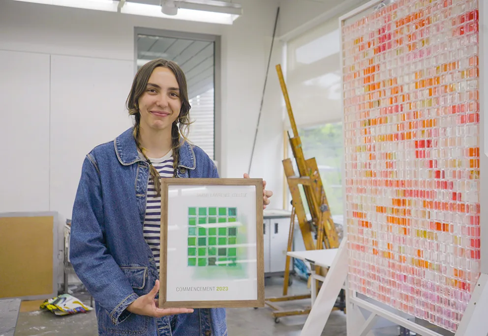 Person holding a framed artwork stands in a studio with colorful grid art and easels in the background.