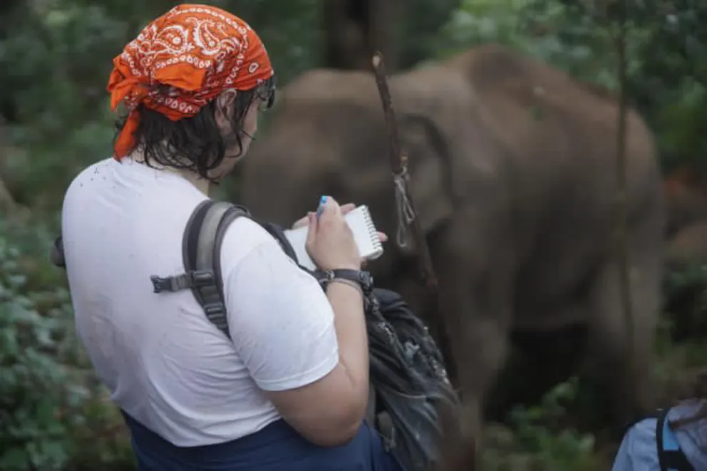 Person with an orange bandana writes in a notebook while standing in a forest with an elephant nearby.