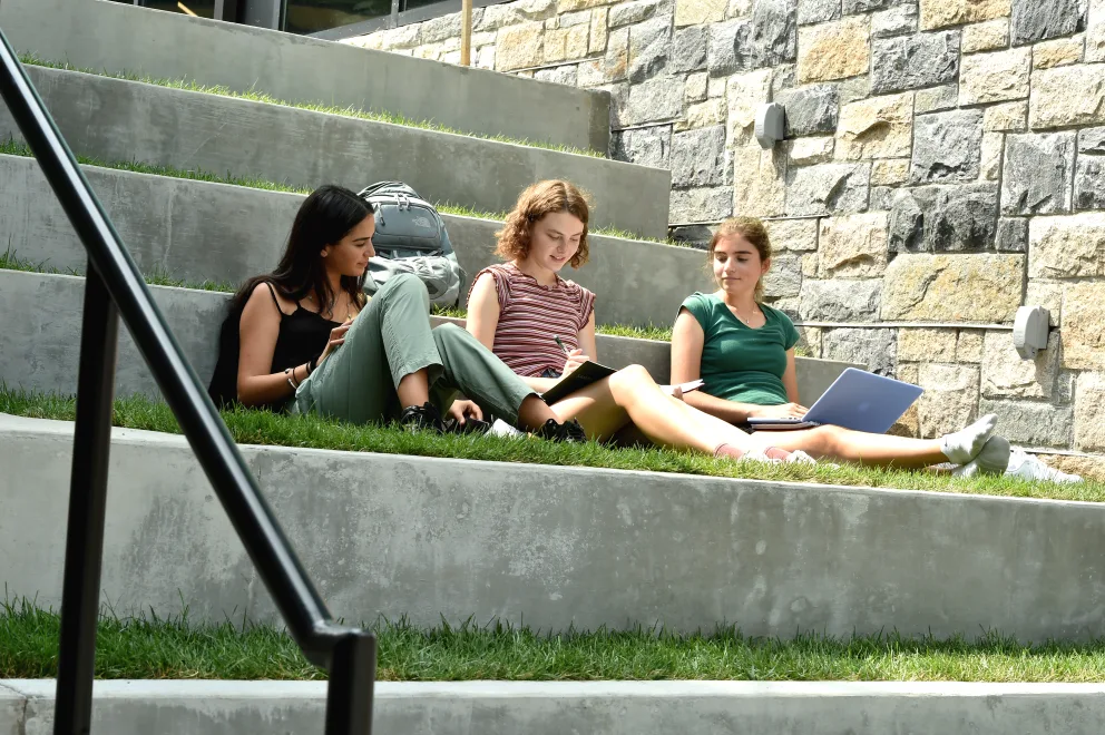 three students are sitting on the grass on the steps outside of a building with their laptops in their hands