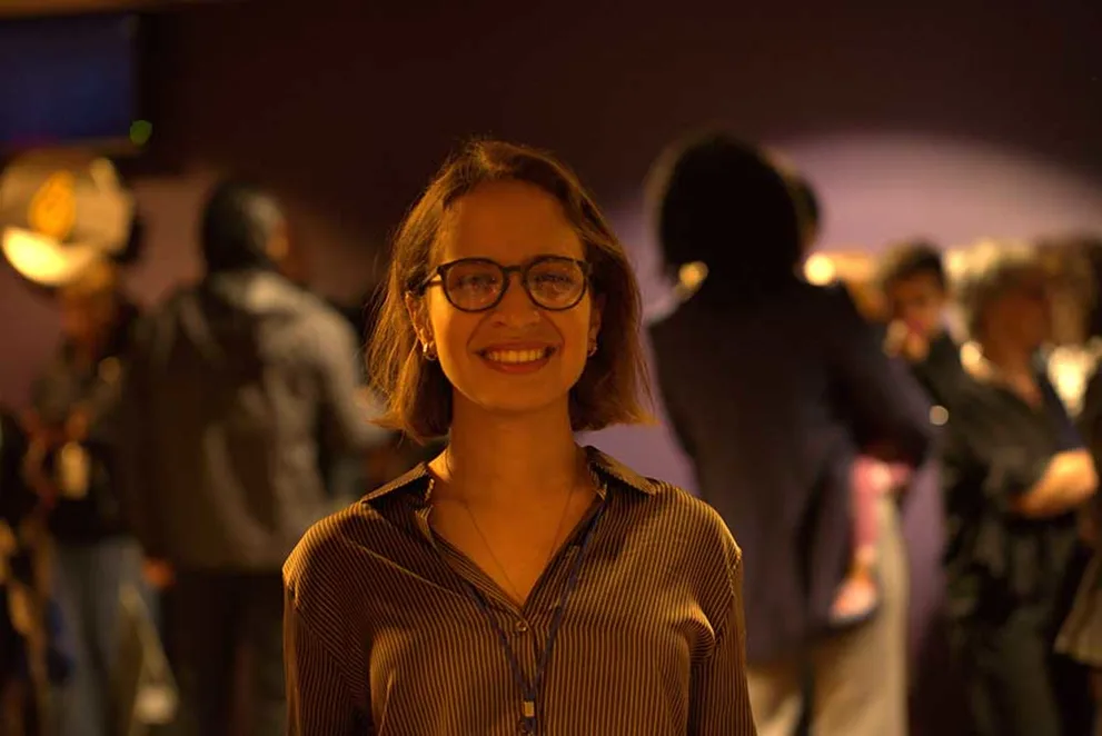 Person with glasses smiling, stands in focus at an indoor event with blurred crowd in the background.