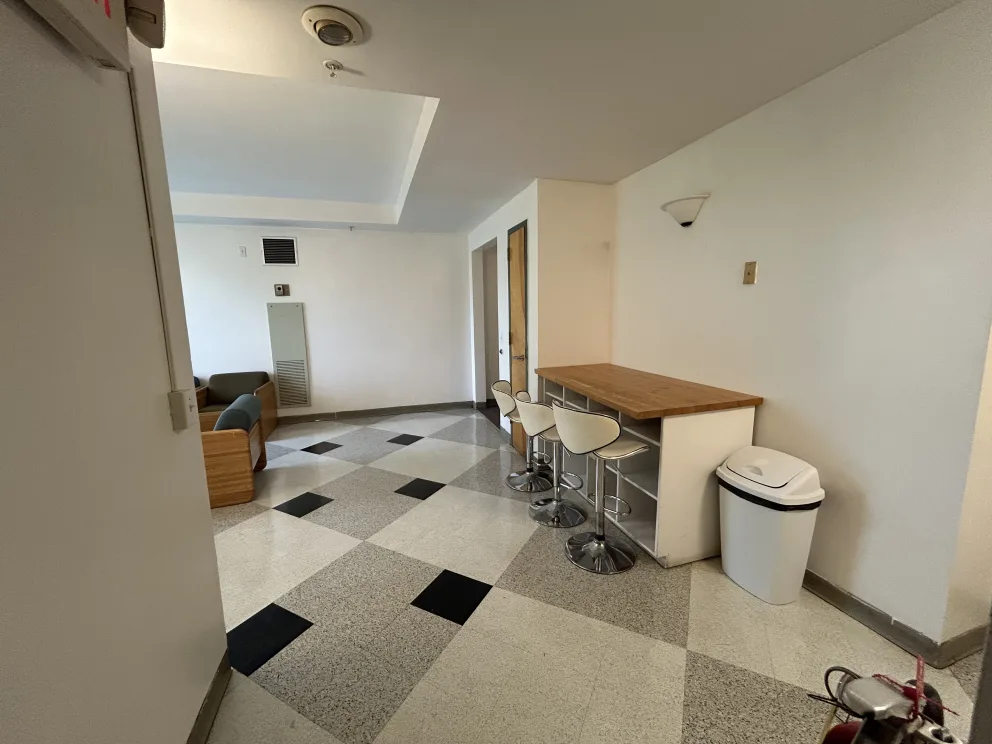 Room with tiled floor, countertop with stools, trash bin, and sofa against the wall. Walls are white, and natural light enters from unseen windows.