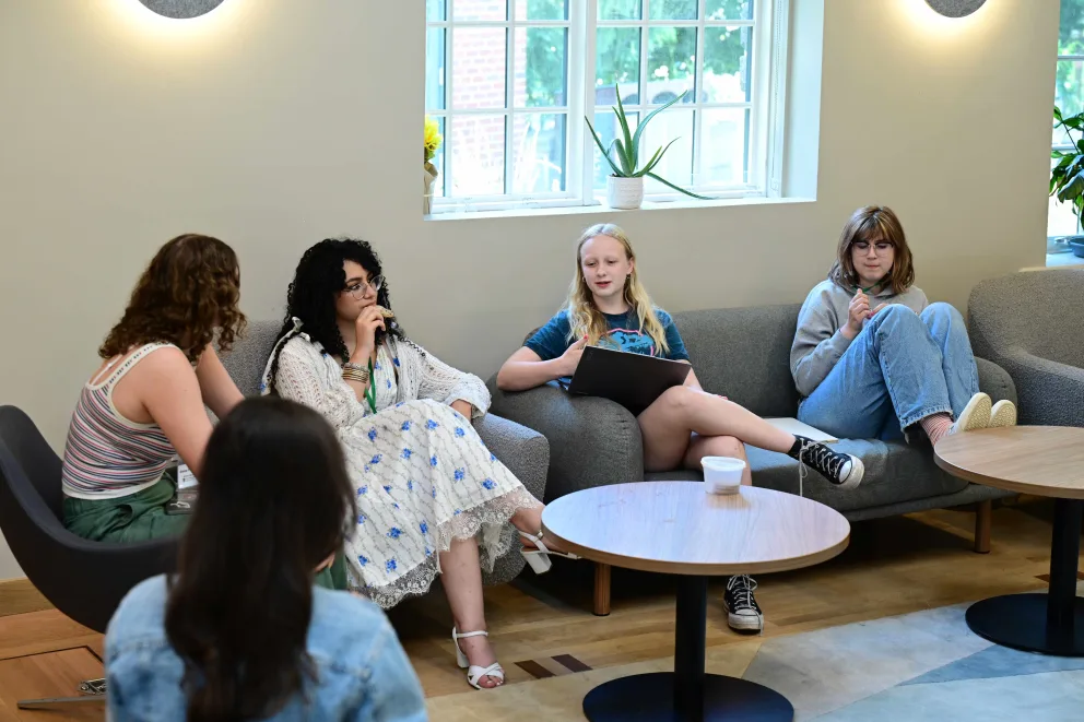 Four people sit in a brightly lit lounge, engaged in conversation. They are seated on couches and chairs around small tables, with a window in the background.