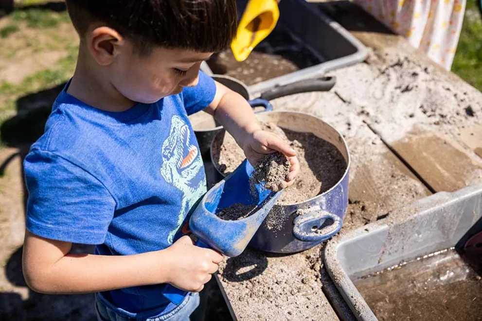 Child in blue shirt scoops dirt into a pot with a plastic shovel while playing outside.