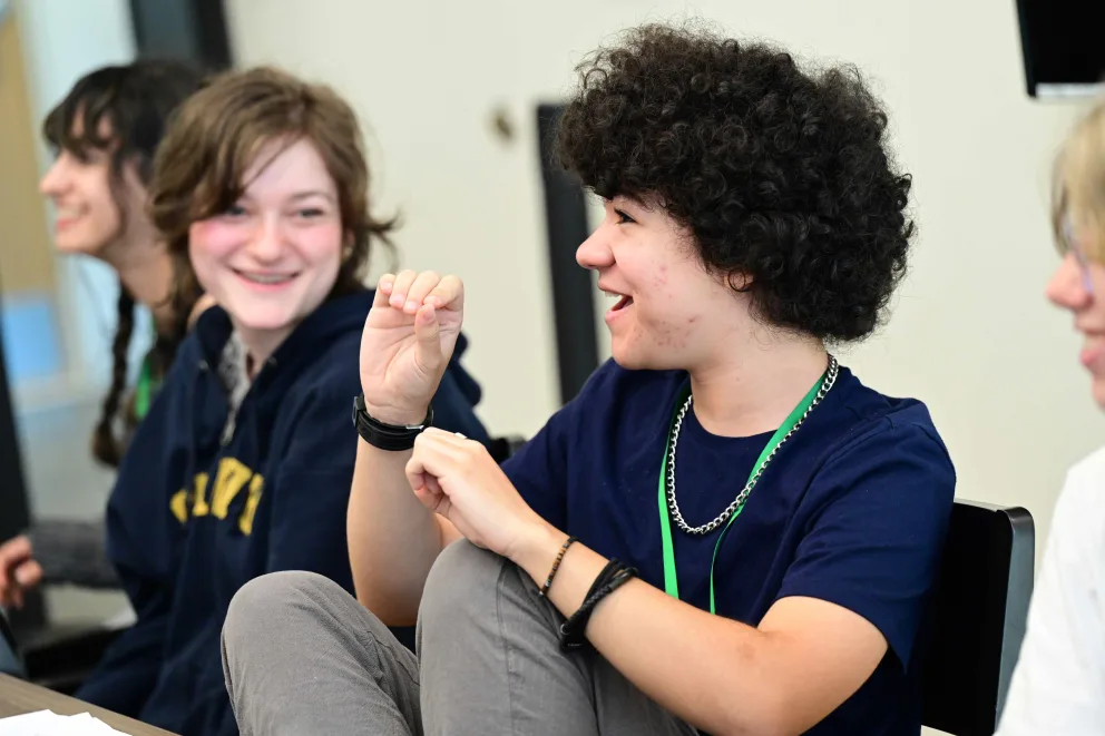 Three people sitting at a table, smiling and engaging in conversation, with one person gesturing with their hand.