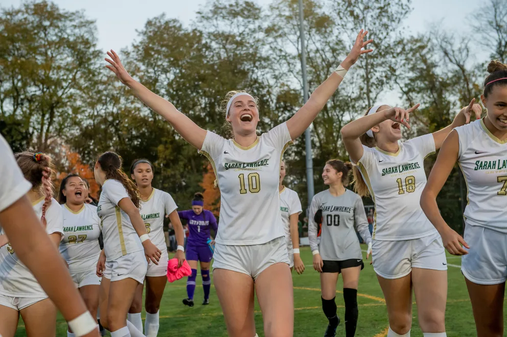 Soccer players in white uniforms celebrate on a field with raised arms and smiles, surrounded by trees.