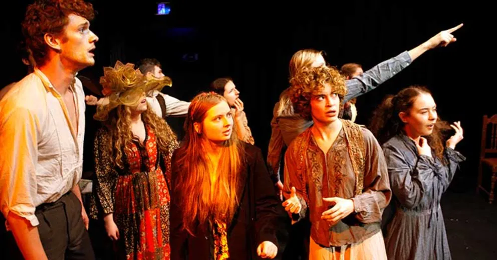 A group of performers on stage in dramatic poses, wearing period costumes, with some looking and pointing towards something in the distance.