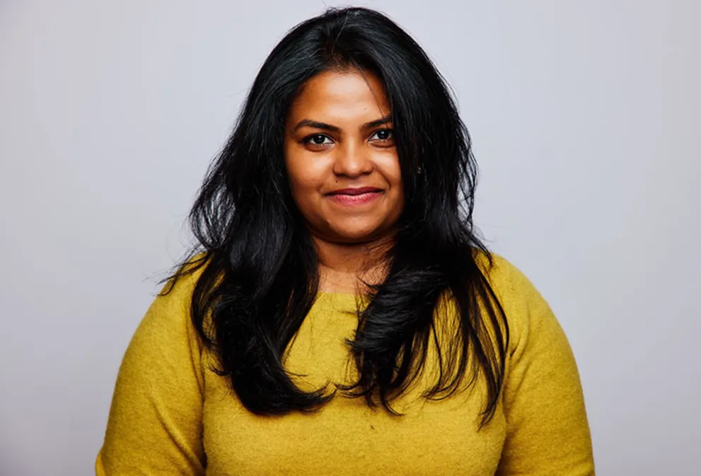 Person with long dark hair wearing a mustard sweater, smiling softly against a plain light background.