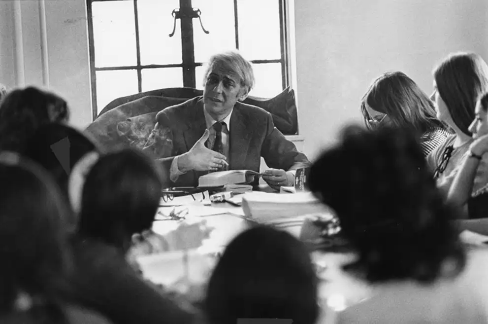 A person in a suit speaks at a table surrounded by others taking notes, with a window in the background.