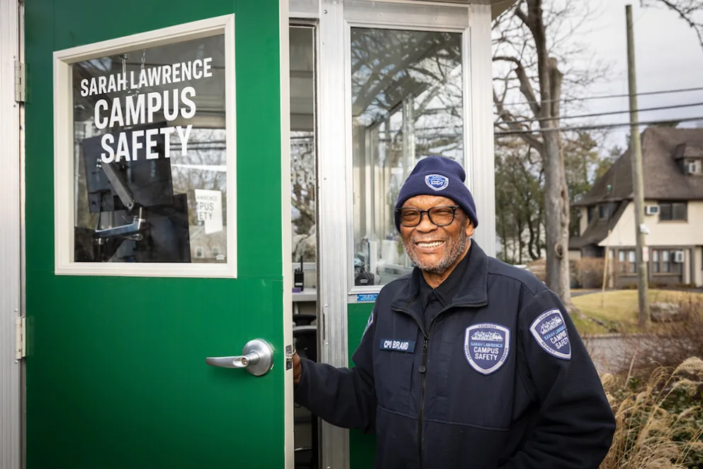 Person in campus safety uniform smiling and standing by a green door labeled "Sarah Lawrence Campus Safety," with houses in the background.