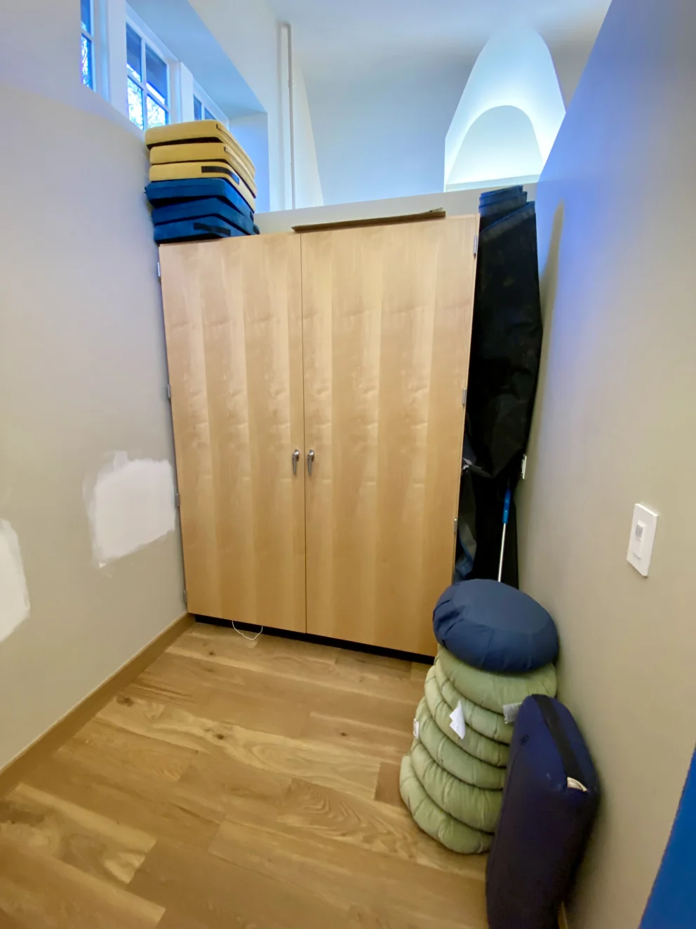 Wooden cabinet in corner with foam mats on top, and stacked cushions beside it.