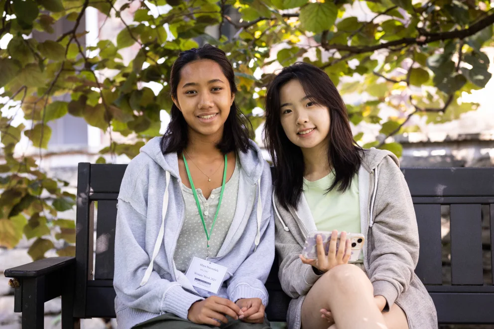 Two people sitting on a bench, one holding a phone. They are outdoors with leafy branches in the background.
