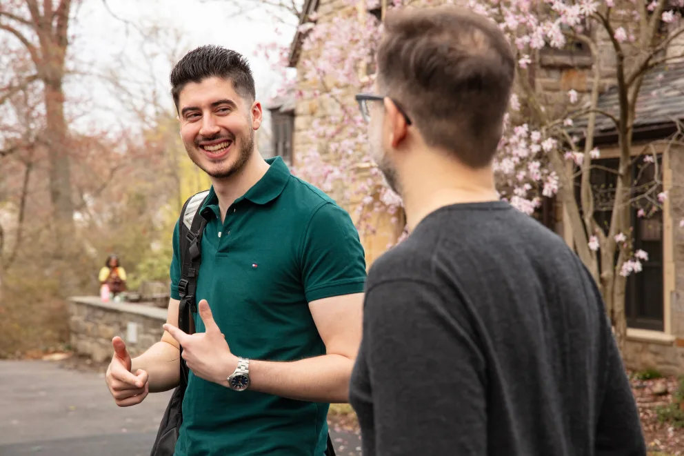 Two people chatting and smiling outdoors near a building with blooming trees. One person gestures with thumbs up.