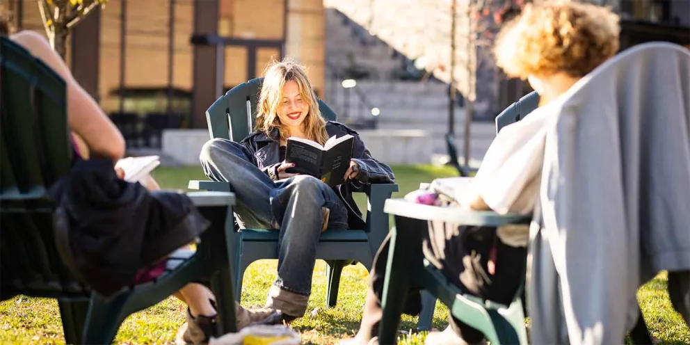 Person reading a book, seated in a circle of chairs with others on a sunny lawn. Buildings and stairs in the background.