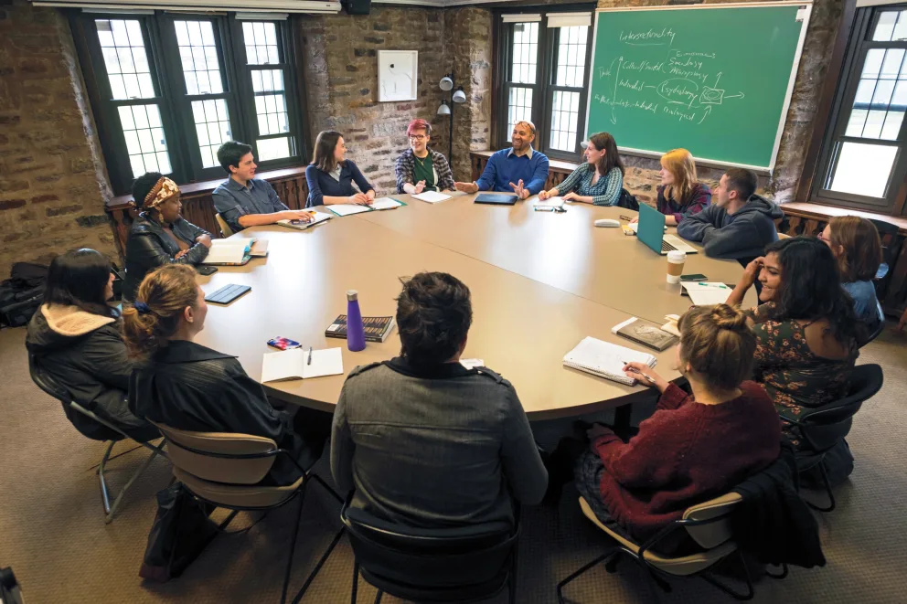 Group of people sitting around a large round table in a classroom, with a chalkboard and windows in the background.