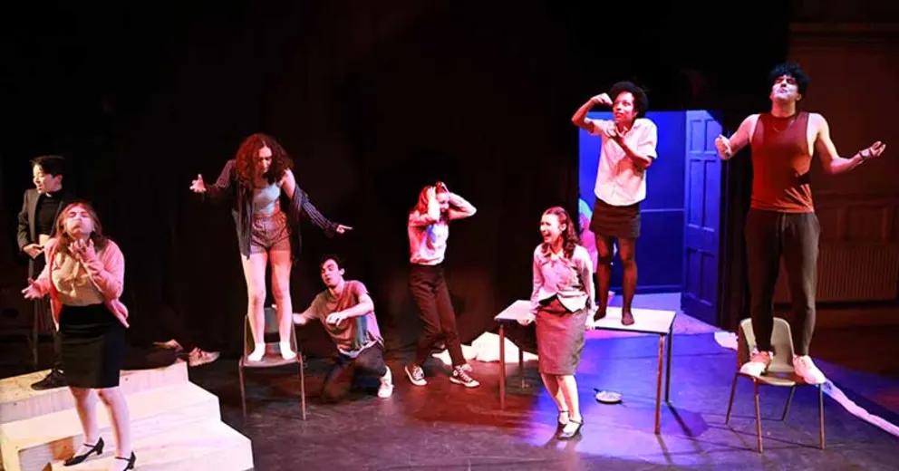 Performers on stage express varied emotions, standing and gesturing dramatically under colorful lighting. The background is dark with a blue door.
