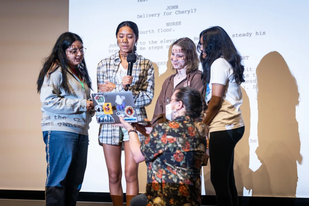 Five people collaborating on a project, gathered around a laptop with a microphone. Text is projected on a screen behind them.