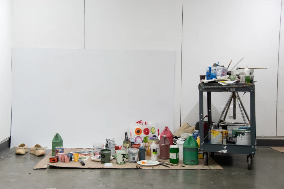Art studio with canvas, various paint cans, brushes, and supplies scattered on the floor and a cart against a white wall.