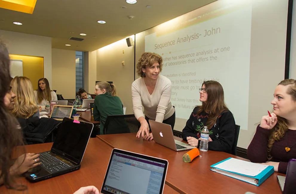 Person standing and speaking to students seated at tables with laptops in a classroom. A slide titled "Sequence Analysis" is projected on the wall.