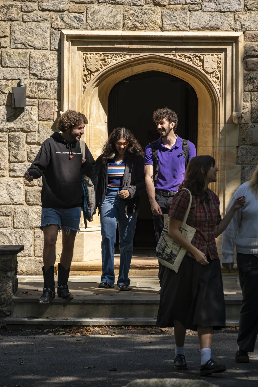 Three people exit a stone building, talking and smiling. Two others walk by in the foreground, with one holding a tote bag.