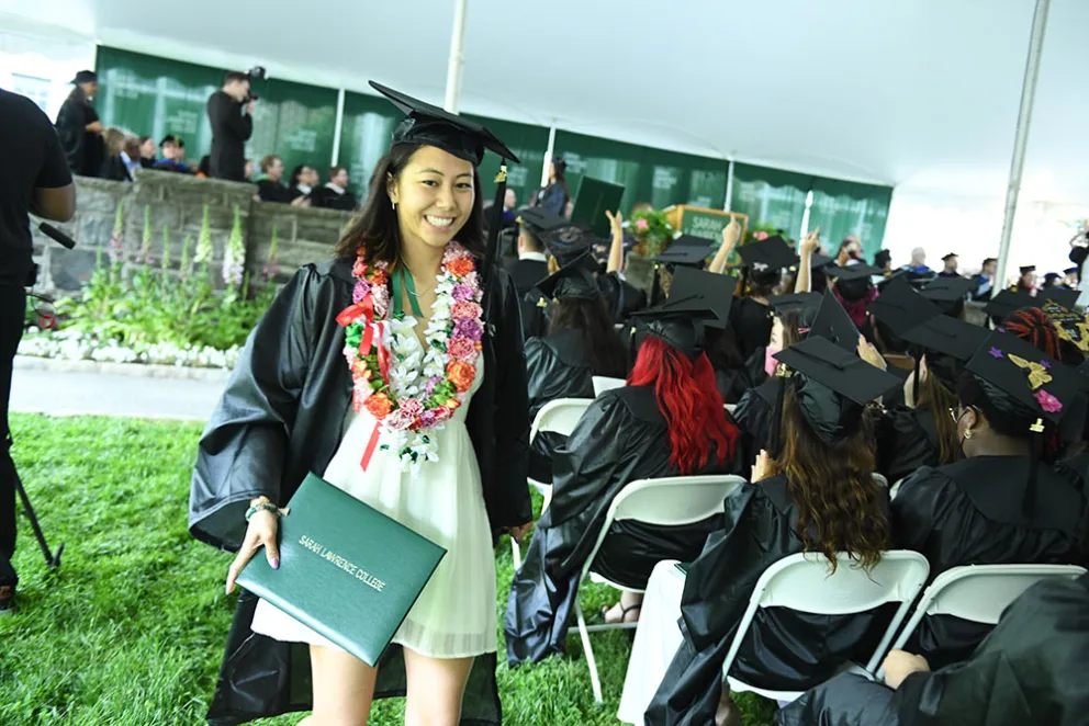 Graduate in cap and gown holds diploma and smiles while walking past seated graduates under a tent during a ceremony.