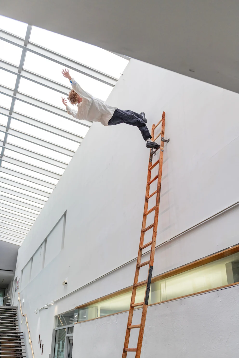 Person in mid-air jumps from a ladder in a modern, well-lit room with a high ceiling and large windows.