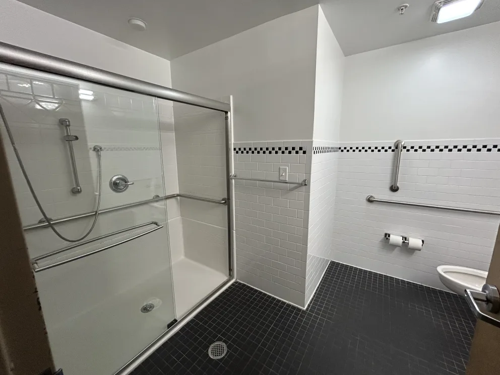 Accessible bathroom with a roll-in shower, grab bars, black and white tiled walls, and a wall-mounted toilet with a toilet paper holder.