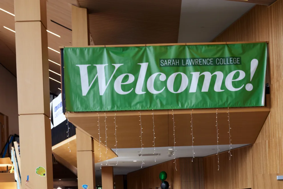 Green banner with "Sarah Lawrence College Welcome!" text, hanging indoors. Wood-paneled walls and string lights are visible.