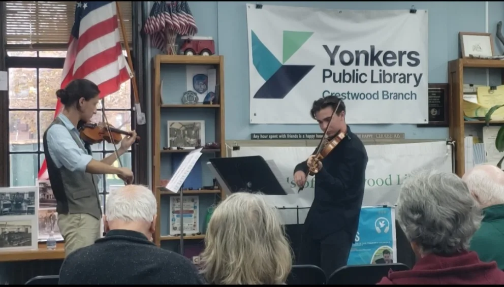 Two individuals playing violins in a library setting, with an audience watching. An American flag and library sign are visible in the background.