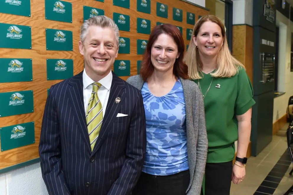 Three people smiling and posing in front of a wall with green and yellow logos.