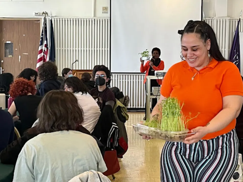 A woman is standing in a room with a box of food, holding a box of green vegetables, and smiling while surrounded by a group of people.
