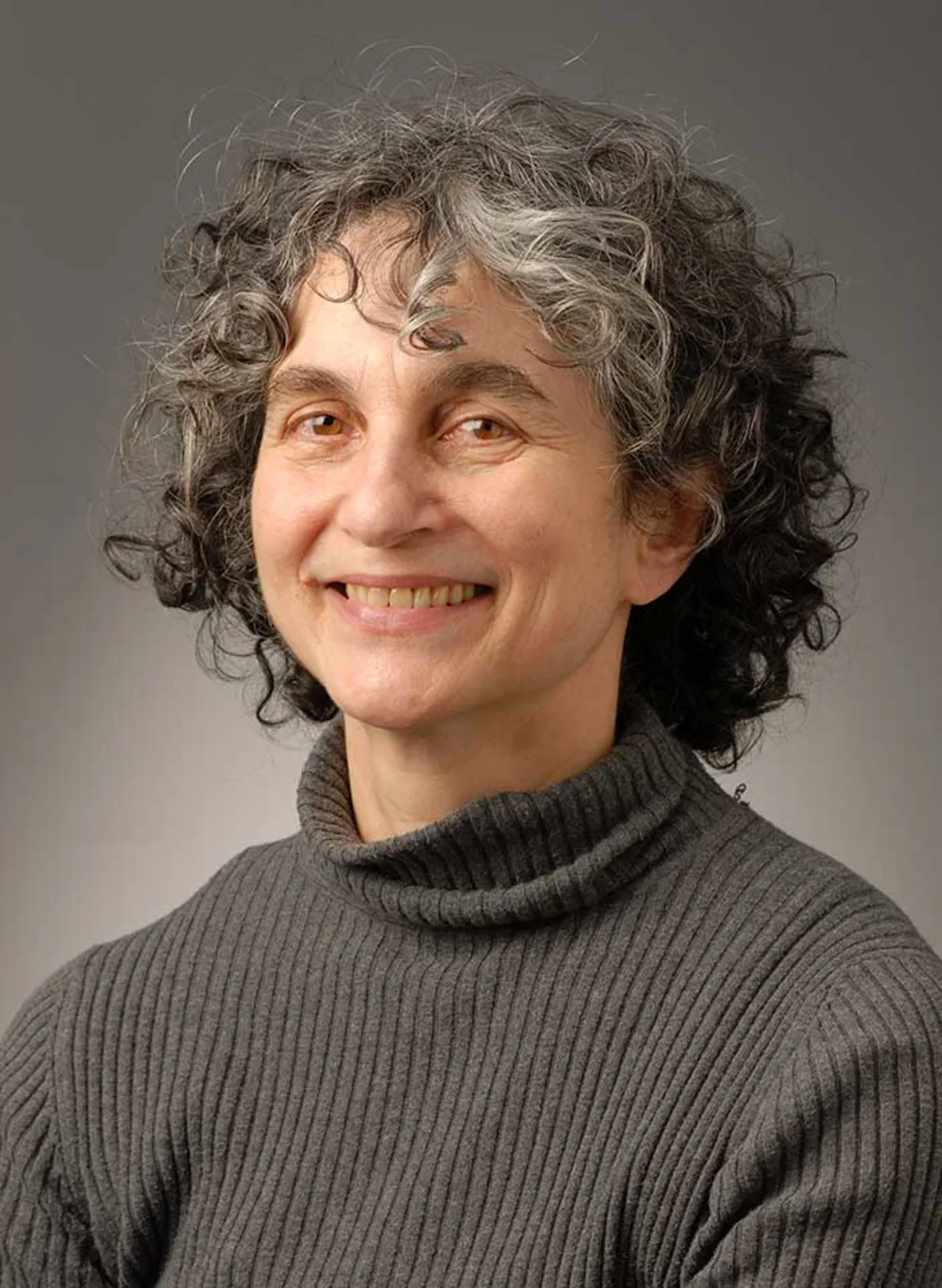 Person with curly gray hair, smiling, wearing a gray turtleneck sweater against a plain background.