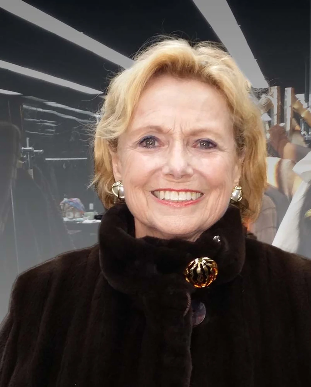 Person smiling, wearing a dark fur-collared coat with large button and earrings, indoors with blurred background.