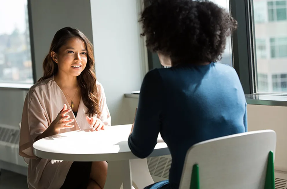 Two people sitting at a round table talking in a brightly lit room with large windows.