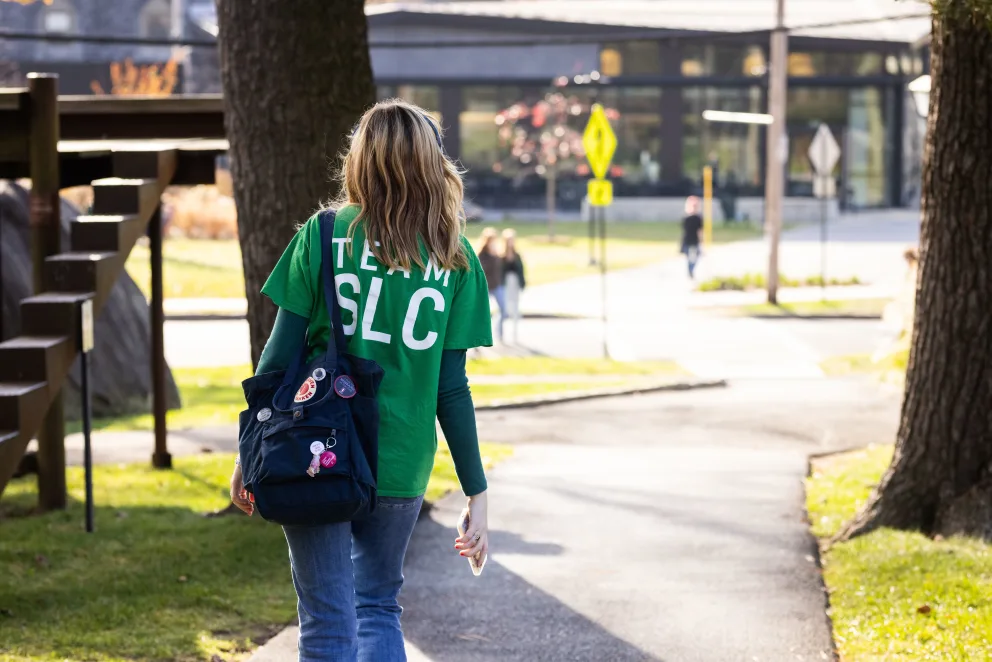 Person in a green "Team SLC" shirt walks on a path with trees and buildings in the background, carrying a decorated bag.
