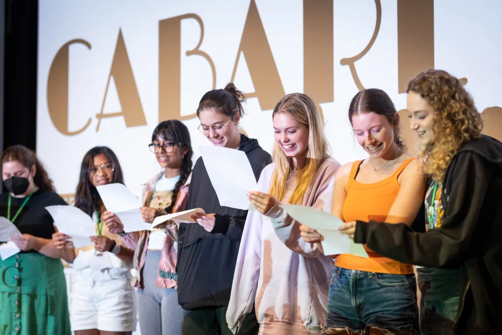 Seven people standing and holding scripts, smiling, with "CABARET" partially visible on a screen behind them.