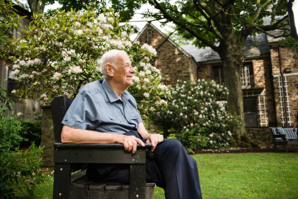 A person with white hair sits on a bench in a garden with flowering bushes and a stone building in the background.