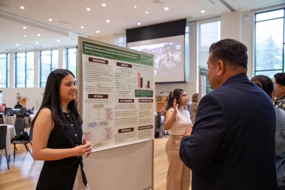 Person presenting research poster to attendees in a conference room with large windows and a screen in the background.