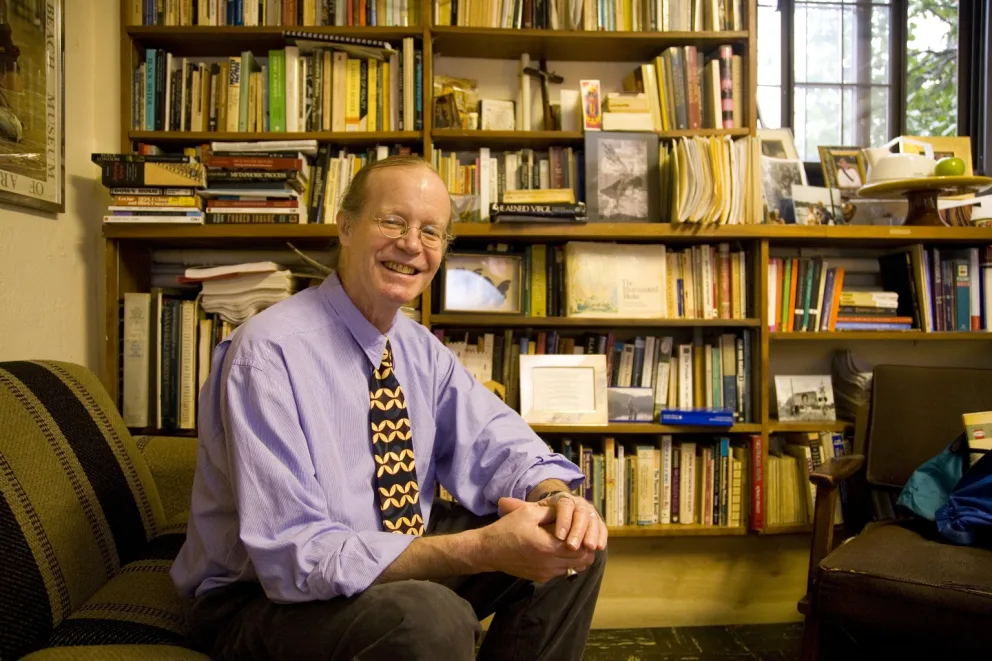 Person smiling, sitting on a couch in an office filled with bookshelves and numerous books in the background.