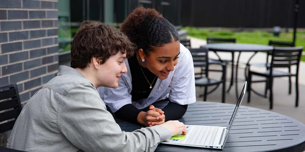 Two people smiling and looking at a laptop screen while sitting at an outdoor table.