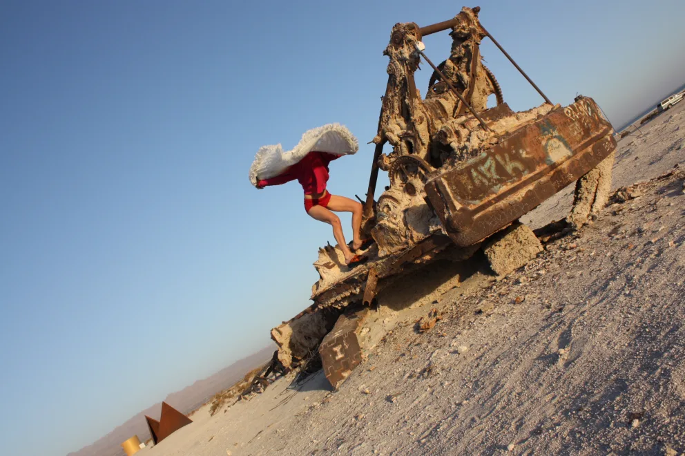 Person in a red outfit leaps from a decaying, rusted machine in a desert landscape under a clear blue sky.