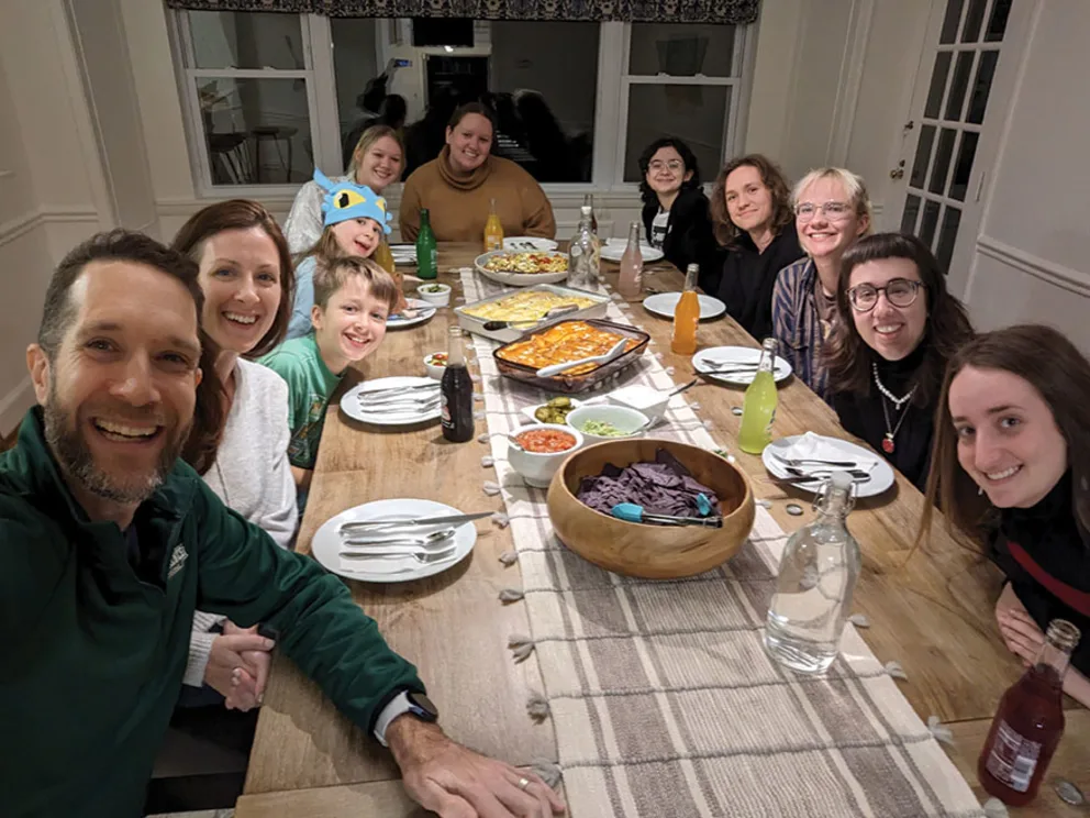 A group of people sitting around a table with plates, drinks, a bowl of chips, and casserole dishes, smiling for a photo in a dining room.