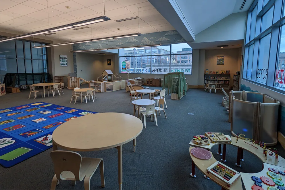 Spacious playroom with small tables, chairs, bookshelves, and colorful play mats. Large windows provide natural light and a view of the cityscape.