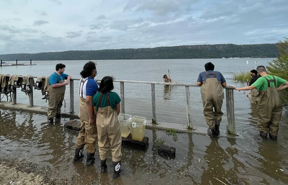 People in waders stand by a railing near a river, observing and working. One person is in the water, surrounded by trees and hills.